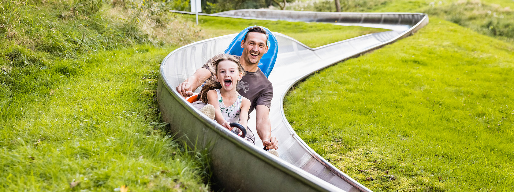 Das Bild zeigt ein Mann und ein Mädchen, die eine Sommerrodelbahn fahren.