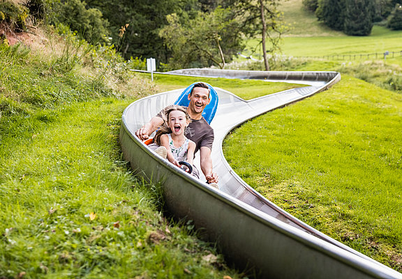 Das Bild zeigt ein Mann und ein Mädchen, die eine Sommerrodelbahn fahren.