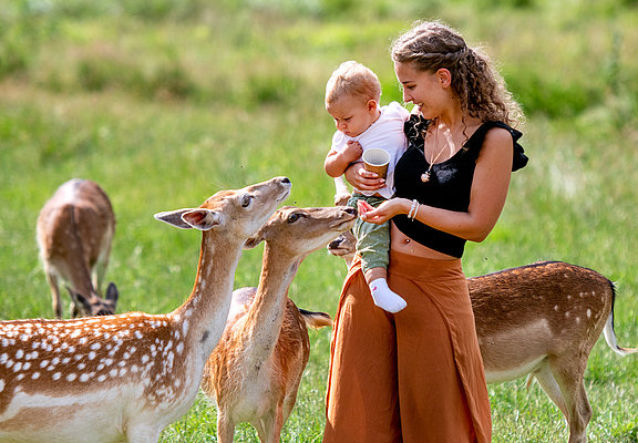 Das Bild zeigt eine Mutter mit ihren kleinen Baby auf dem Arm. Neben ihnen stehen Rehe.