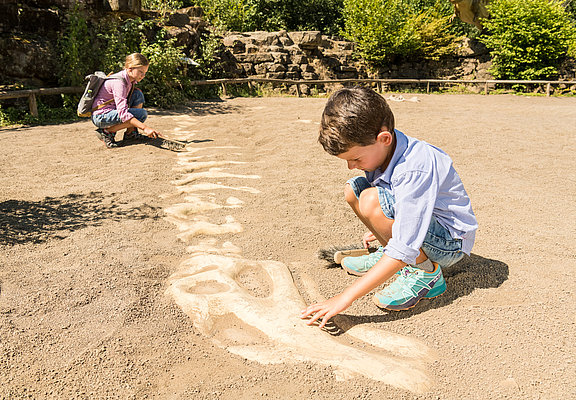Das Bild zeigt zwei Kinder im Dinopark Teufelsschlucht, die auf einer Sandfläche ein Dinoskelett ausgraben.