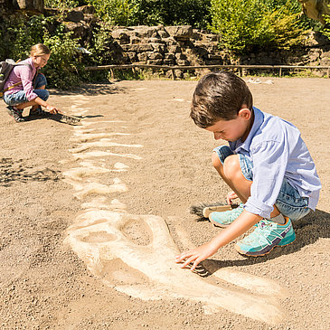 Das Bild zeigt zwei Kinder im Dinopark Teufelsschlucht, die auf einer Sandfläche ein Dinoskelett ausgraben.